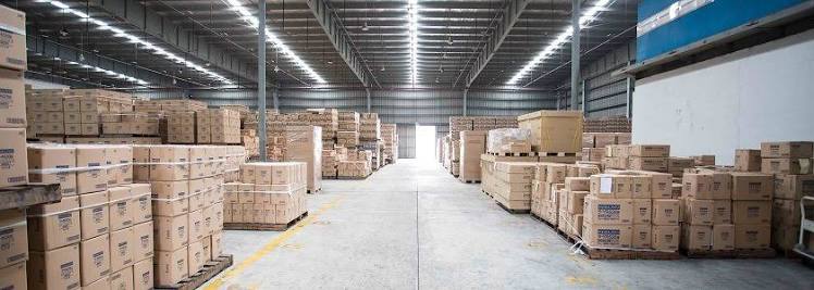 A wide view of a large, modern warehouse in Guangzhou with pallets of cardboard boxes stacked on both sides of a long aisle under bright industrial lighting.