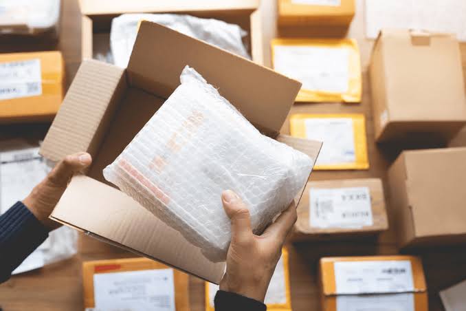 A person's hands opening a cardboard box to inspect a bubble-wrapped item, with other shipping boxes stacked in the background, representing the 2-step dropshipping process from China.
